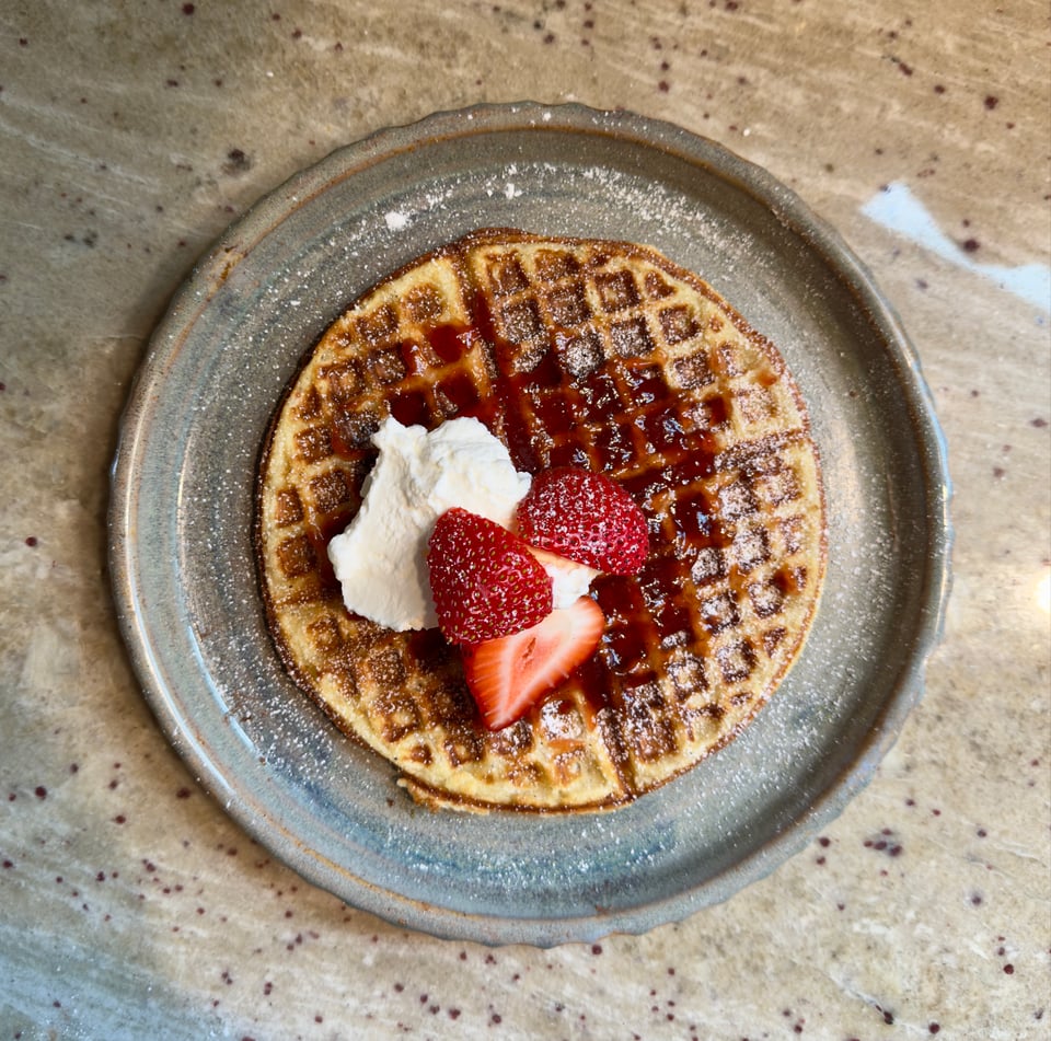 Waffle made with keffir on a plate topped with strawberry jam, strawberries, whipped cream, and powdered sugar.