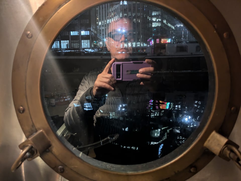 Looking through a brass porthole at night, modern buildings are reflected in the water. My image is a ghostly reflection holding my phone and taking the picture.