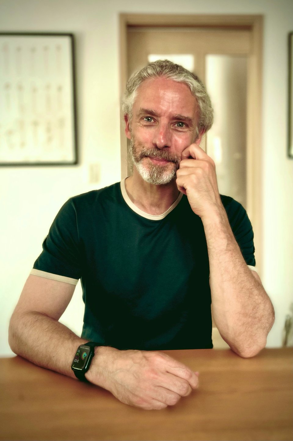 portrait-mode photo of a white man in a green ringer T-shirt sitting at a wooden table, his head resting against his left hand, while his right forearm is on the table. He's smiling, but wishes he'd blotted his forehead a little before the picture
