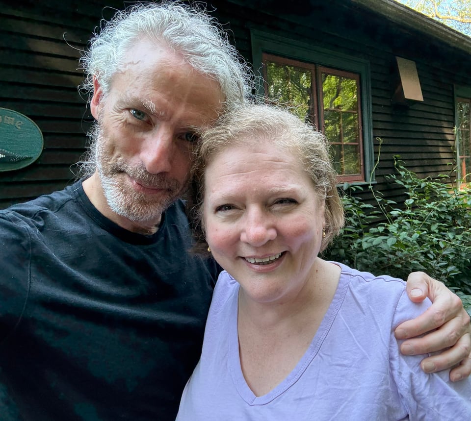 photo of white man and woman standing in front of a wood-sided house, smiling at the camera