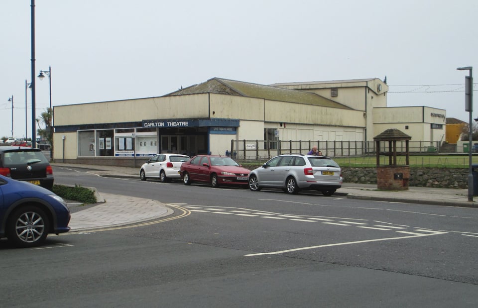 A low squared off theatre building with a higher block at the back. At the front is a slight canopy with the words Carlton Theatre in a rounded blocky font. On the side wall there are indentations that look like old windows.