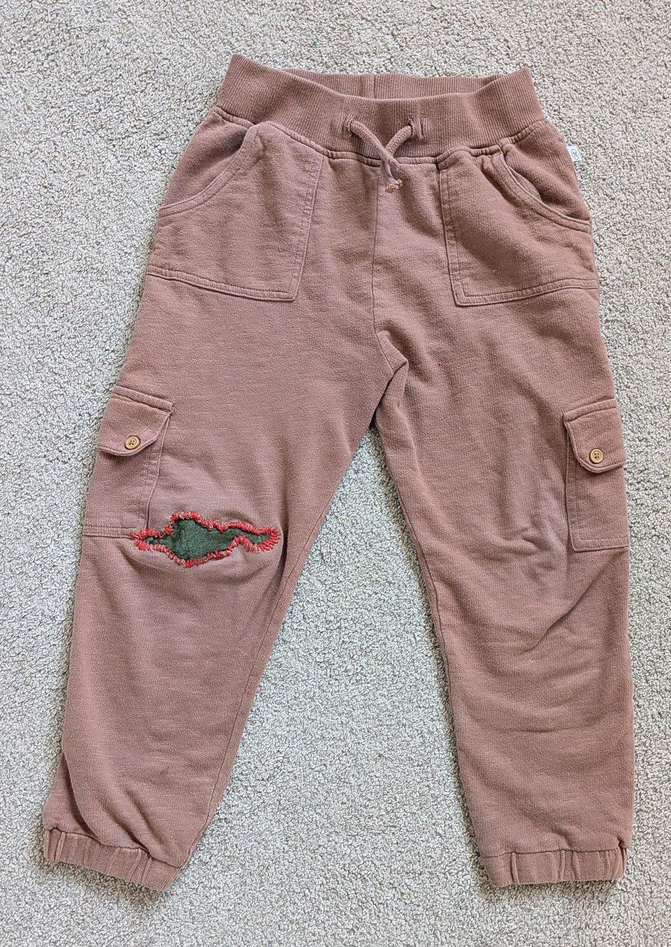 a child's light brown pants have an irregular patch across one knee, made from green fabric with red embroidery floss along the edges. The patch looks almost like a landmass or perhaps a dinosaur.