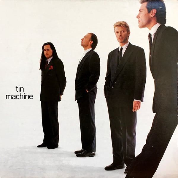 A photograph of the four members of Tin Machine, three white men and David Bowie, who is an alien. They are all dressed in spiffy suits and ties. They are lined up facing to the left, with one member closer to the camera and the last one furthest away from the camera. David Bowie and the guy furthest away from the camera, who has very long brown hair, are looking at the camera while the other two seem to be looking at something to the left and up out of frame.