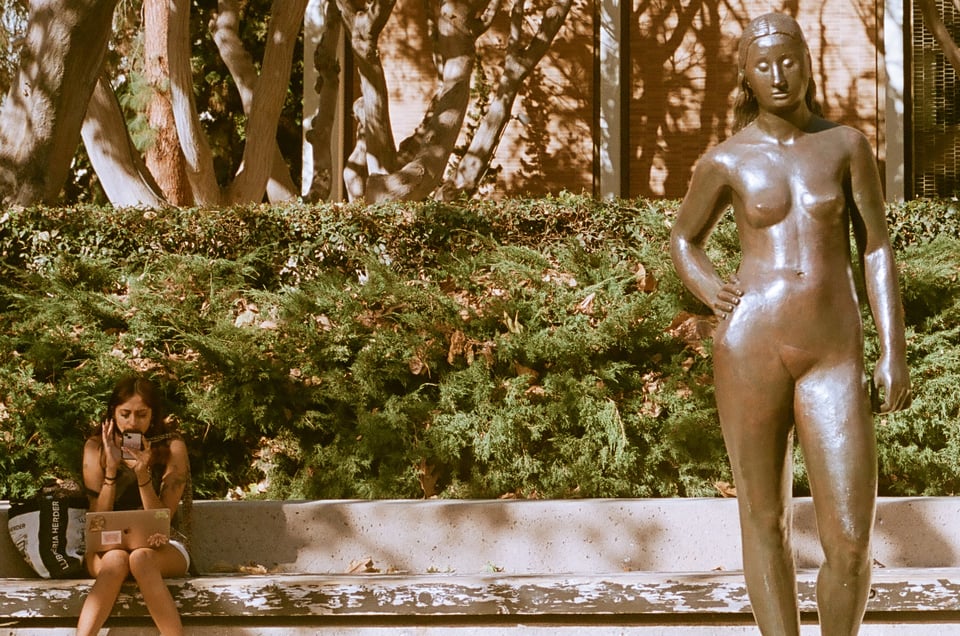 A woman sits on a stone bench and talks on her phone to the left of a bronze cast statue of a nude woman with her hand on her hip