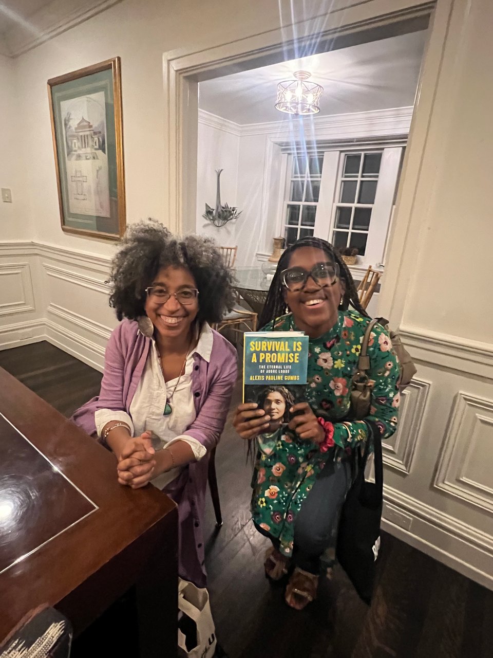 a photograph of two black women, one with glasses and an afro and another with long braids who is holding up a book entitled "Survival is a Promise".
