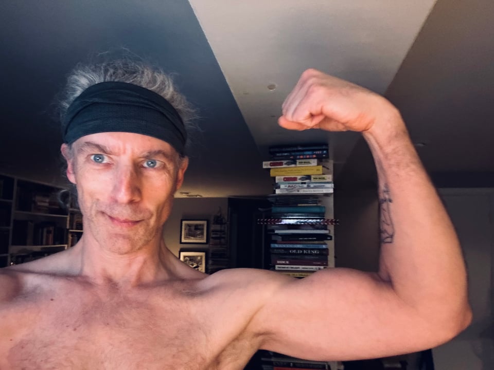 Photo of shirtless white man with dark grey headband flexing his bicep. There's a vertical bookshelf behind him.