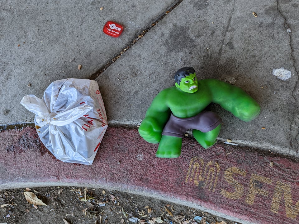 An incredible hulk toy sits on the sidewalk next to a bag of trash. He looks pissed off about it.