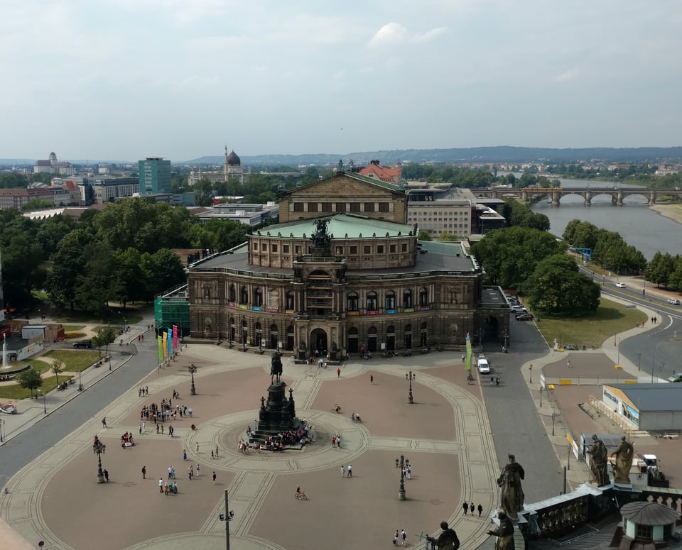 The Dresden Semperoper, a 4-story building in the Neoclassical style, sits across a pedestrian square with a statue in the middle.