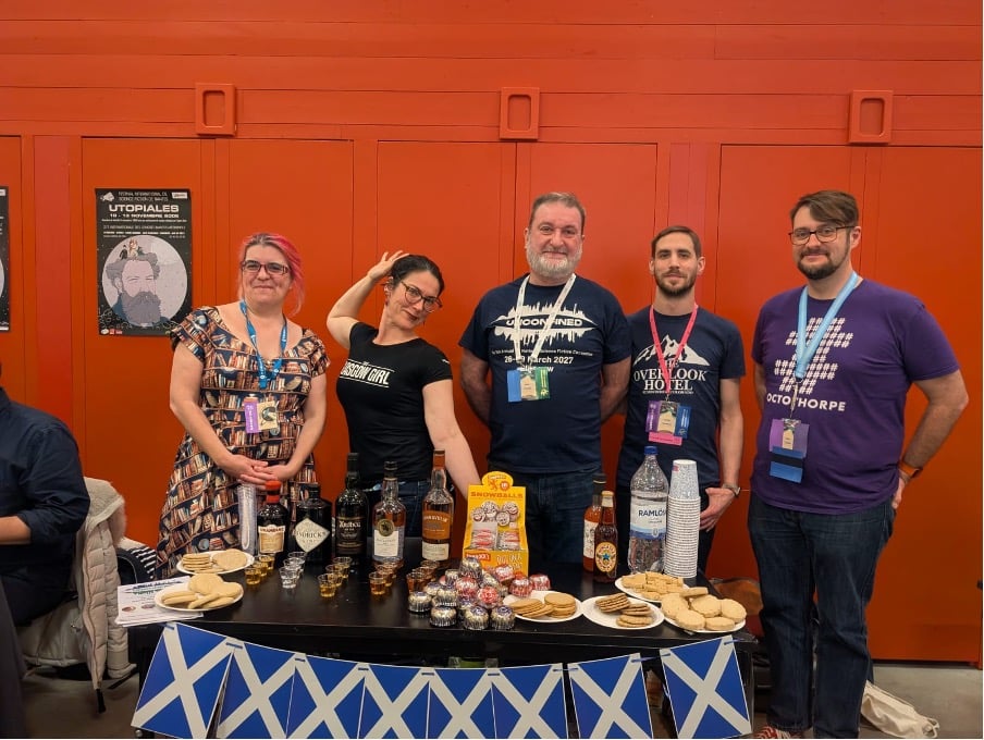 A photograph of five people - two women and three men - standing behind a table with drinks and biscuits on it, the table wrapped in small scottish flags