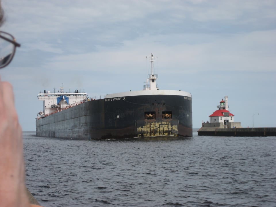A really big (and wide) freighter sailing past a harbor light.