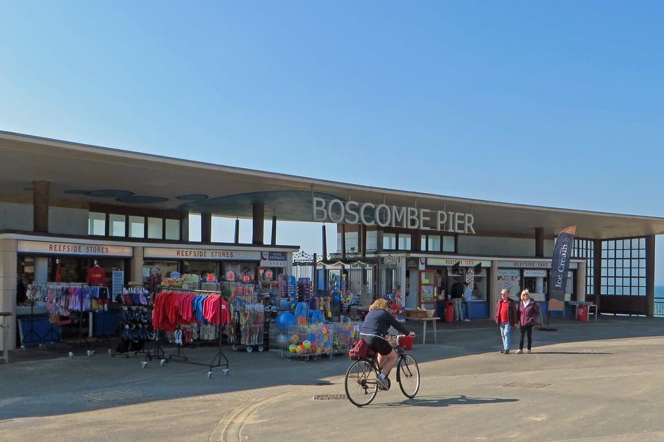 The pier entrance, as described in the main post. It's a sunny day and the shops have racks of seaside things to buy like beachballs and postcards. the cafe is open. Someone is cycling past. The lettering that hangs from the roof is popping nicely.