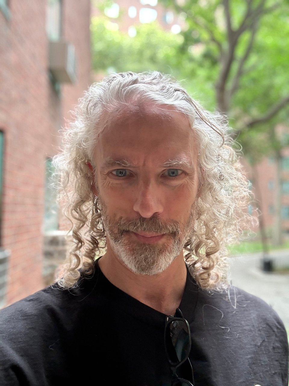 outdoor selfie of middle-aged white man with long, curly hair and a somewhat neatly trimmed white beard; he's wearing a black T-shirt and had a pair of Wayfarers hanging from his color