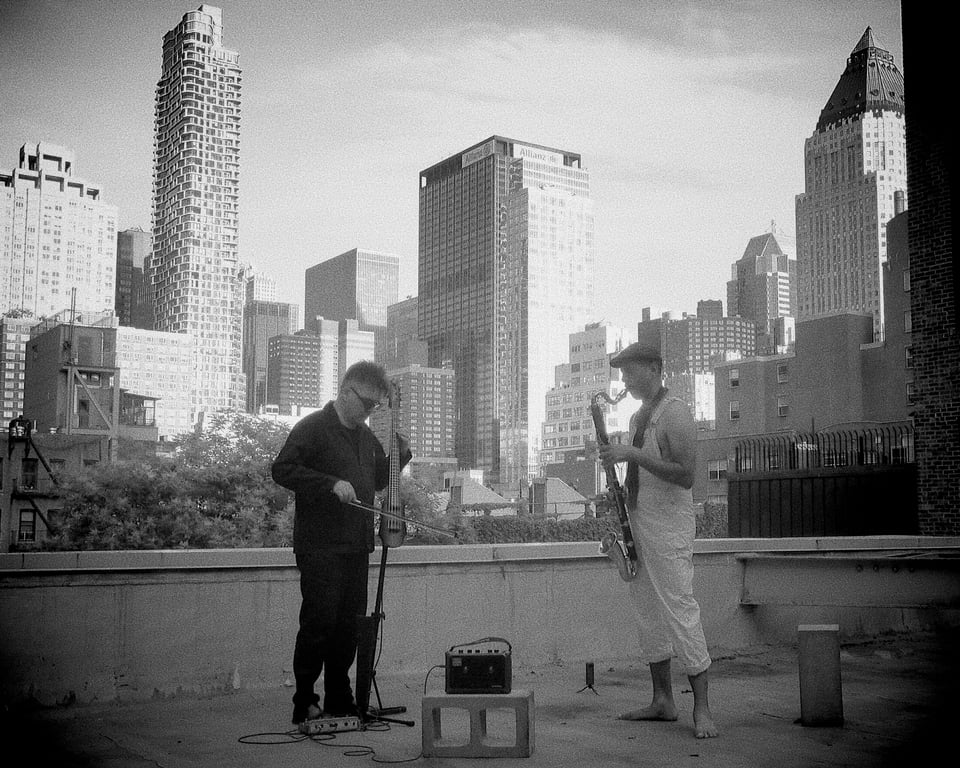 Blake Leyh & Tony Jarvis playing electric cello and bass clarinet in a black and white photo on a Manhattan rooftop with towering buildings behind them