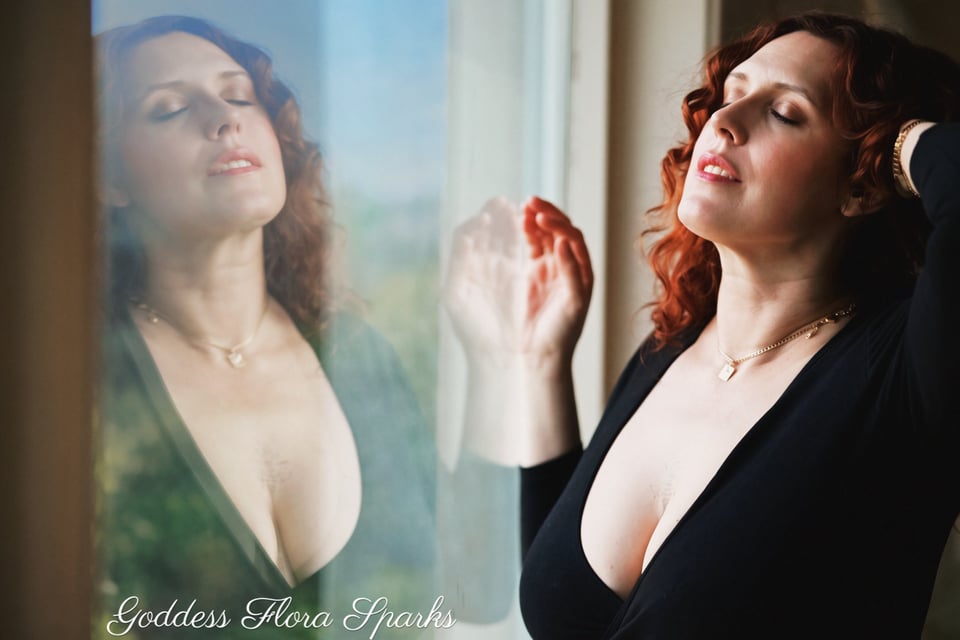 flora with her flora lifted and hand in her hair, left hand on a window. We can see her reflection in the window