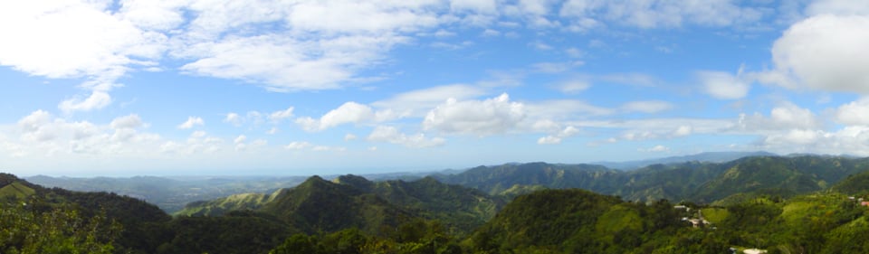 Expansive view of green mountains under a bright blue sky with scattered fluffy clouds.