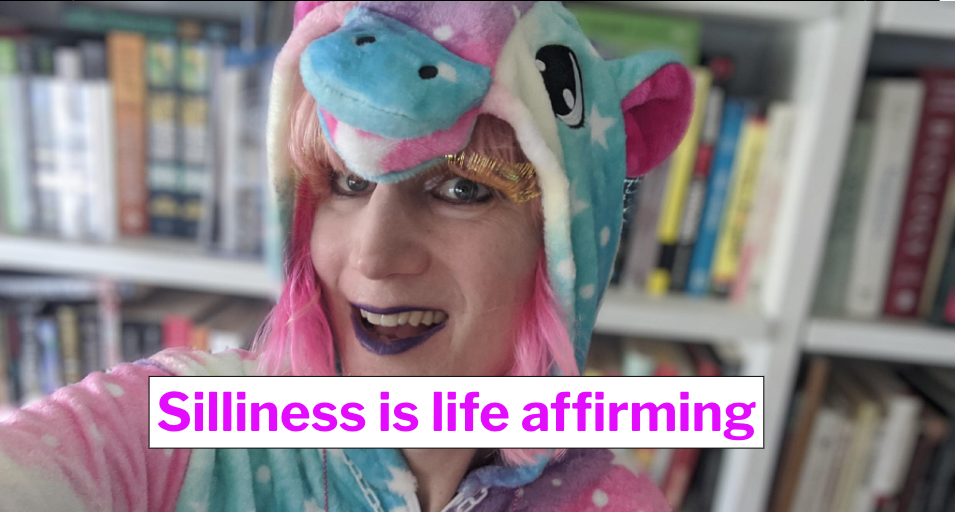 Me wearing a cartoon unicorn onesie with my pink hair poking out and a goofy grin on my face, with gold eyelashes. Banner says, SILLINESS IS LIFE AFFIRMING