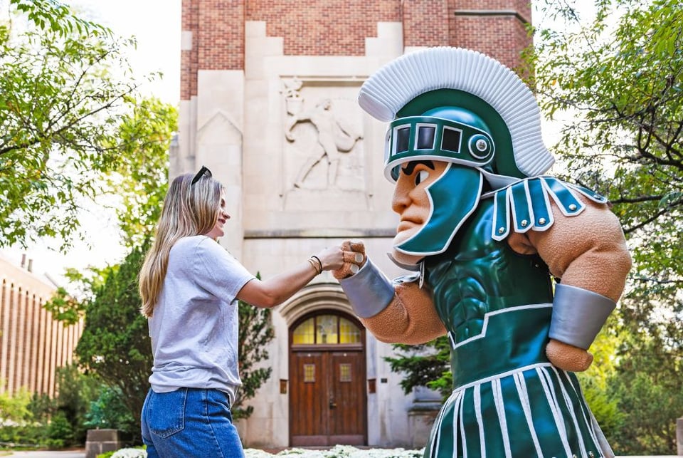A JMC student with long blonde hair is smiling and fist-bumping the Michigan State University Spartan mascot, who wears a green helmet and armor. They are standing outdoors in front of a brick building adorned with a relief sculpture, surrounded by trees.