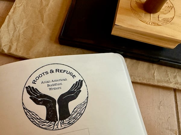 The upper half of the image shows part of a carved, square wooden rubber stamp on top of a rectangular black ink pad on top of a rectangular piece of craft paper. The lower half of the image shows the upper right corner of a white notebook page inked with a black circular stamp with the words “Roots & Refuge Asian American Buddhist Writers” and an image of two hands, palms upturned, in the shape of a bowl. Hair-like roots sprout from the wrist of the two hands. The stamp, ink pad, craft paper, and notebook rest on a wood table.