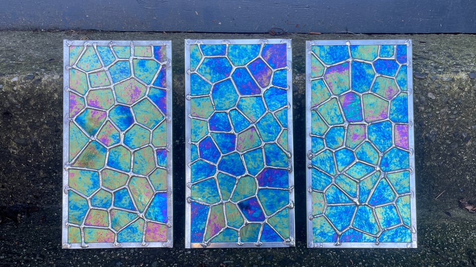 Three rectangular stained glass panels are sitting against a dark grey concrete. The glass pieces are are a brilliantly iridescent in shades of purple and teal and are cut and assembled as an irregular cellular (Voronoi) pattern.