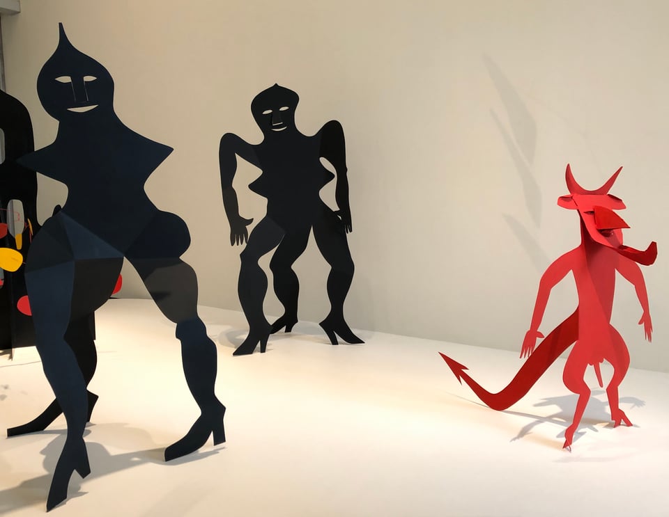 Three two-dimensional cut-out metal and wire sculptures, two black, one red, resembling demons or devils. They stand in a bright and cheery exhibition space, with white walls and flooring.