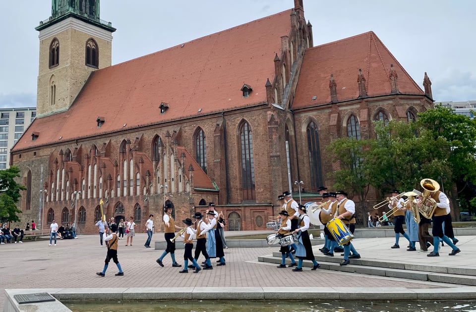 A local civilian marching band marches while playing near the TV tower in Berlin. In the background is St. Marienkirche, a 15th century red brick Gothic church