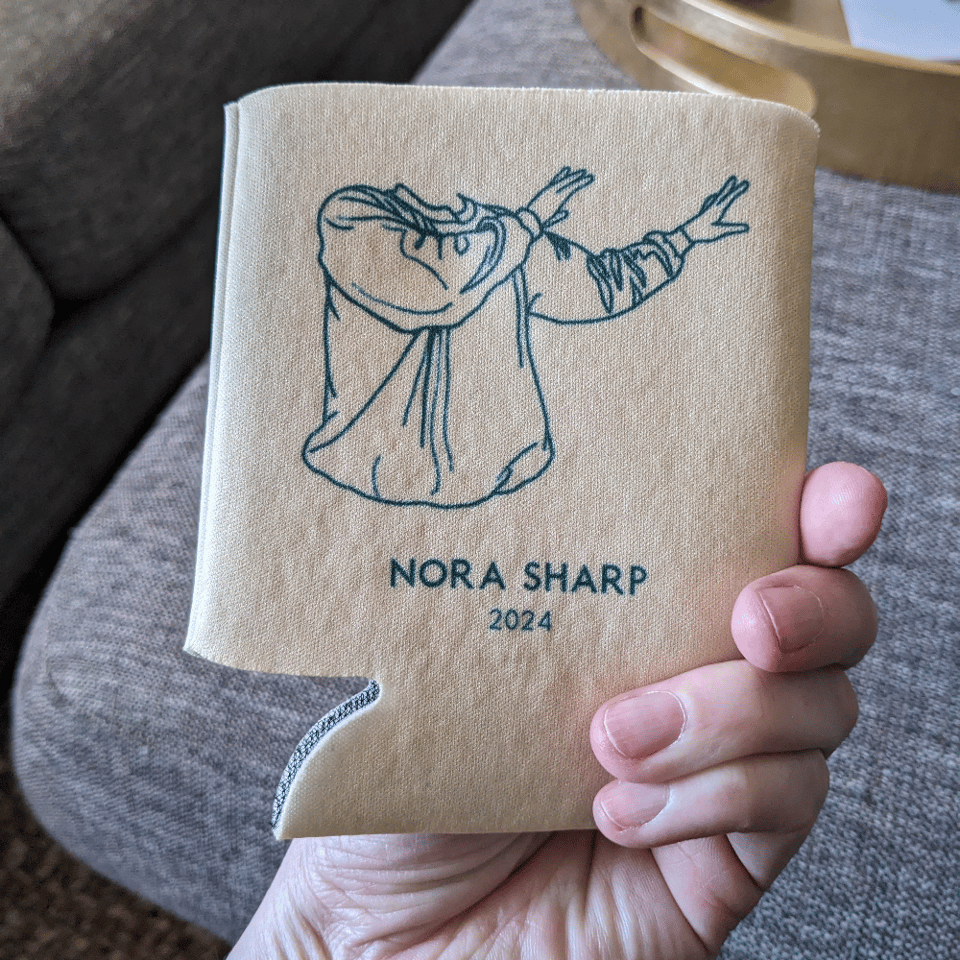 A hand holding a pail beige coozie with evergreen printed image and text. The image is the silhouette of a torso and arms in a sweatshirt making a demonstrative gesture. The text reads Nora Sharp 2024.
