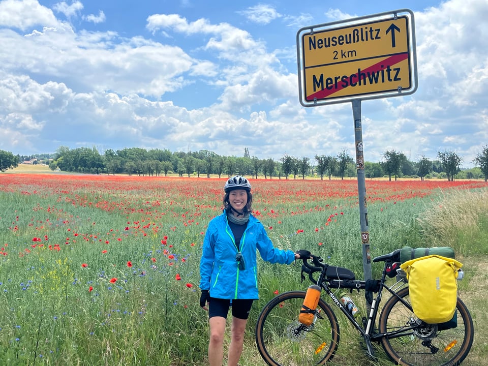 Jenny and her bike standing in front of a field of red poppies, with a sign indicating that Neuseusslitz is 2 km away, and we are exiting Merschwitz.
