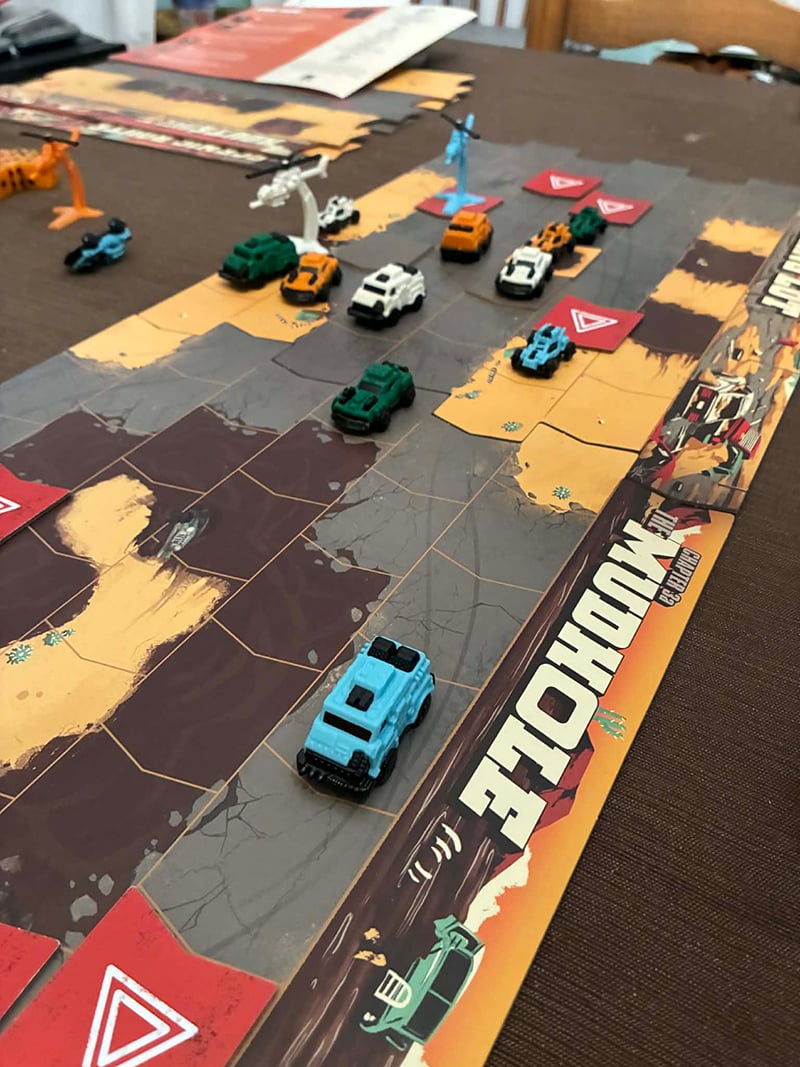 The board for the game Thunder Road: Vendetta is set up on my dining room table. There are multiple pieces of board, forming a road. There are little plastic cars on the road. You move them and try to destroy your opponents' cars.
