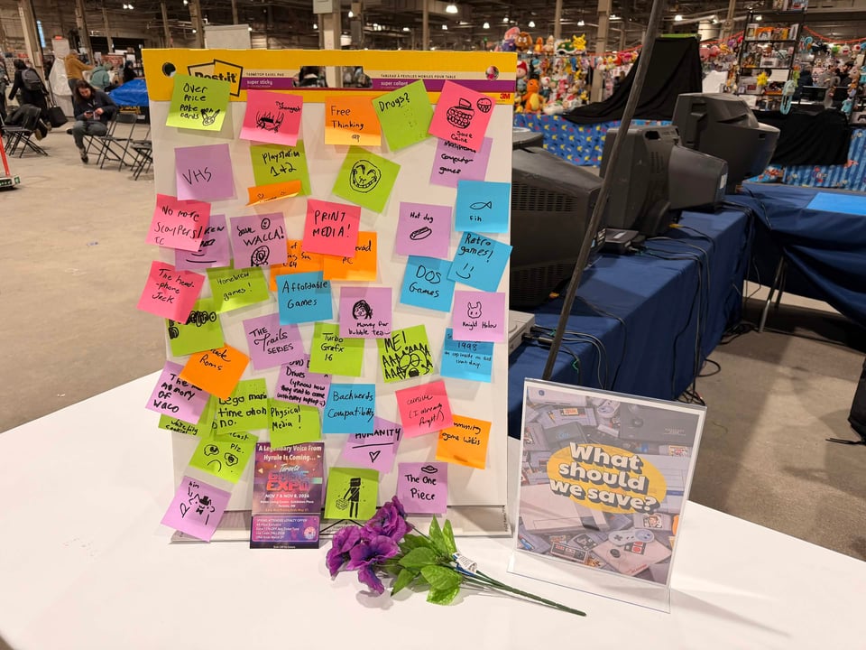 A convention table display featuring a large Post-it easel pad covered in colorful sticky notes with handwritten answers to the prompt "What should we save?" — visible responses include "VHS," "Print Media," "Retro Games," "DOS Games," "Affordable Games," and "Physical Media" — alongside a framed sign reading "What should we save?" and a bouquet of purple artificial flowers on the white tablecloth, with a busy expo hall and vendor booths in the background.