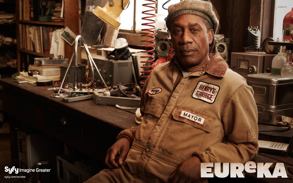 Henry in his workshop wearing a cap and a work jacket that says "HENRY's GARAGE" and "mayor"                       