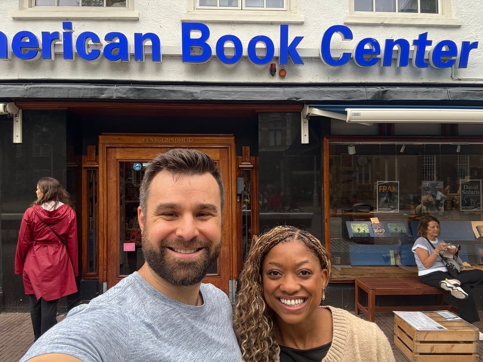 Two friends, Joe and I, smiling and taking a selfie outside the American Book Center in Amsterdam. The storefront has bold blue lettering and a wooden entrance door. Behind us, the window displays books.