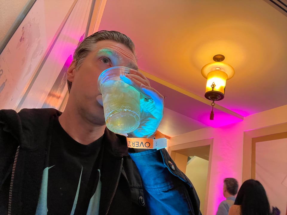 A man sips a mixed drink from a plastic cup in a hallway with a lovely art deco lamp