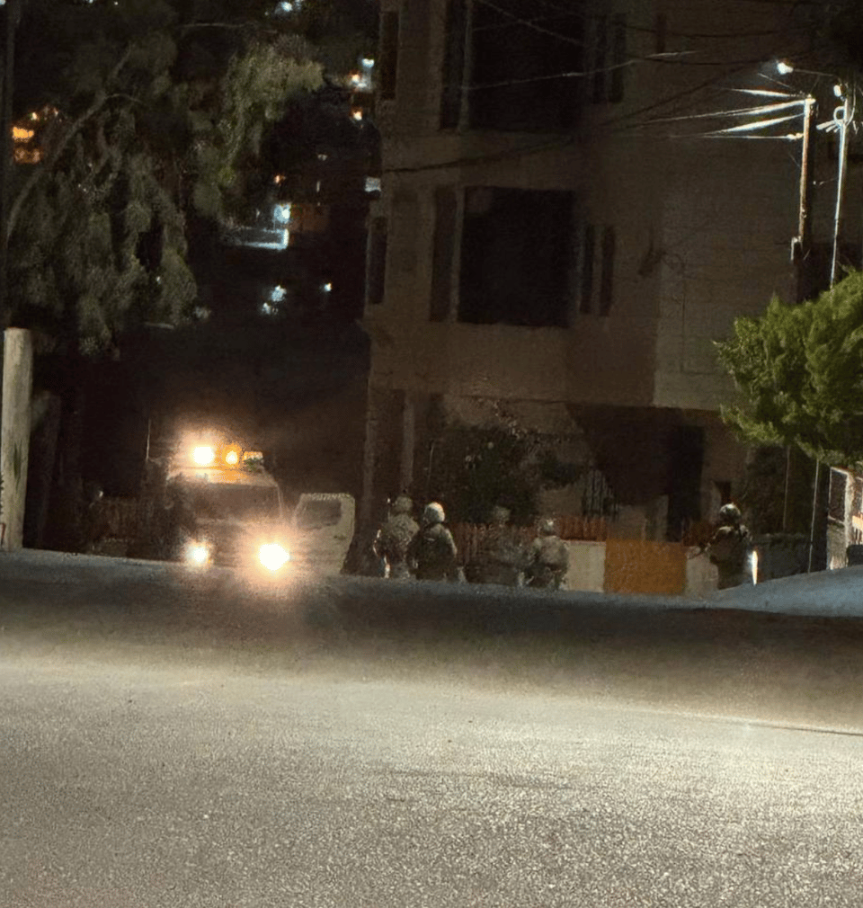 A distance view of a dark streeet with a streetlight by an apartment building with a jeep with lights and six soldiers
