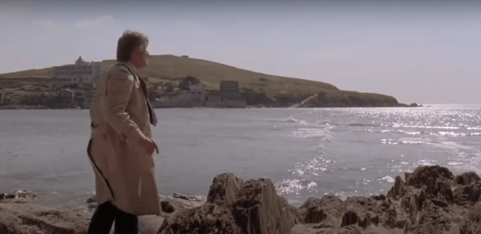 Cain (Kirk Douglas) stands on the mainland in a classic beige trenchcoat, looking in horror at the tide that has come in. Burgh Island and the hotel are in the background.