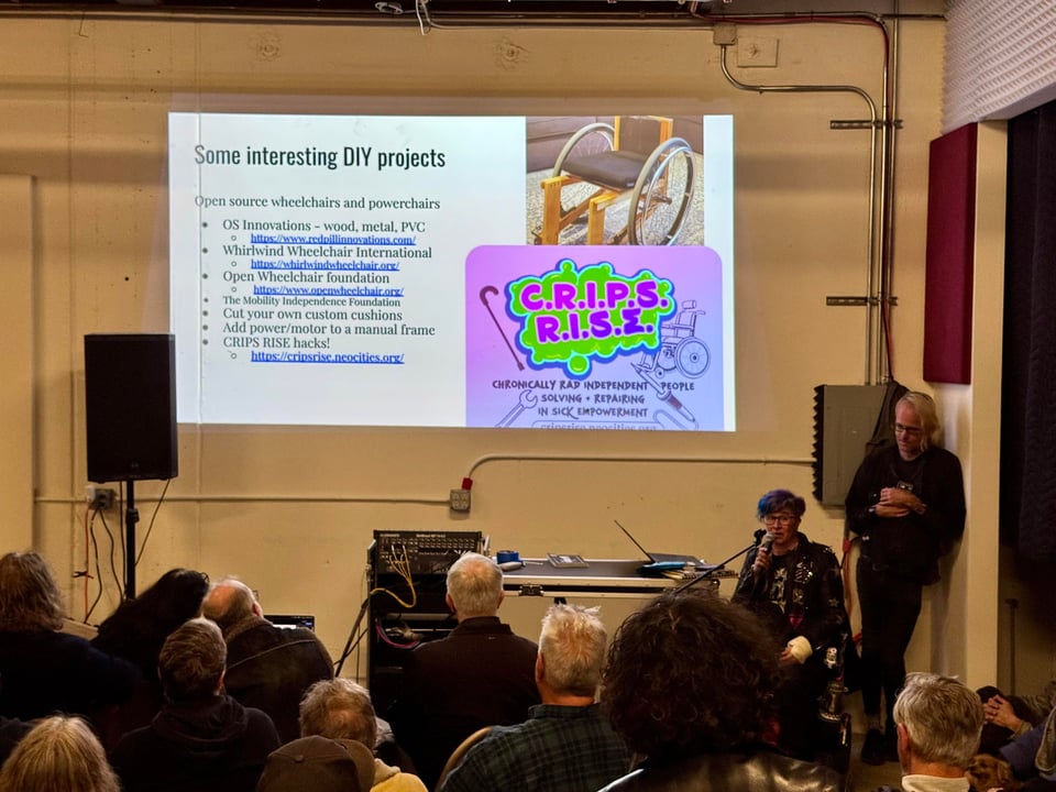 Liz in powerchair speaking into a microphone, slide showing CRIPS RISE graphic and a list of open source wheelchair projects