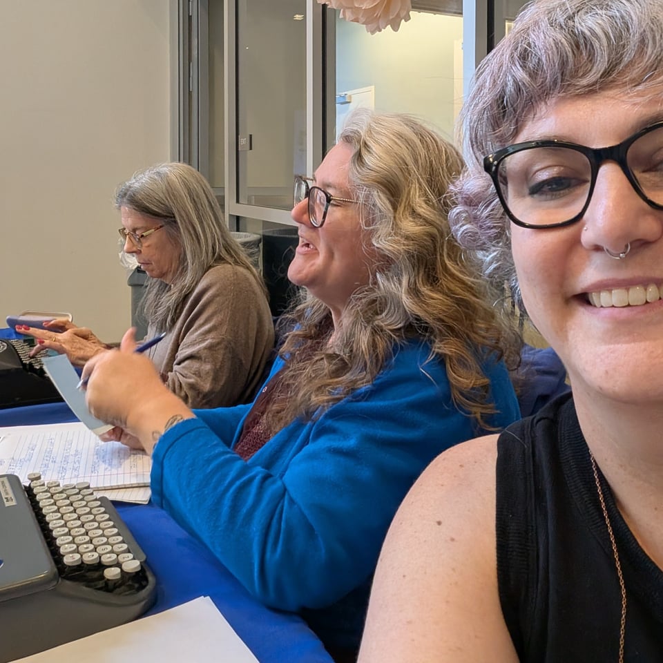 Three poets sit at a table with typewriters. Two are at work and the third, who is taking the selfie of them, is smiling and holding the camera.