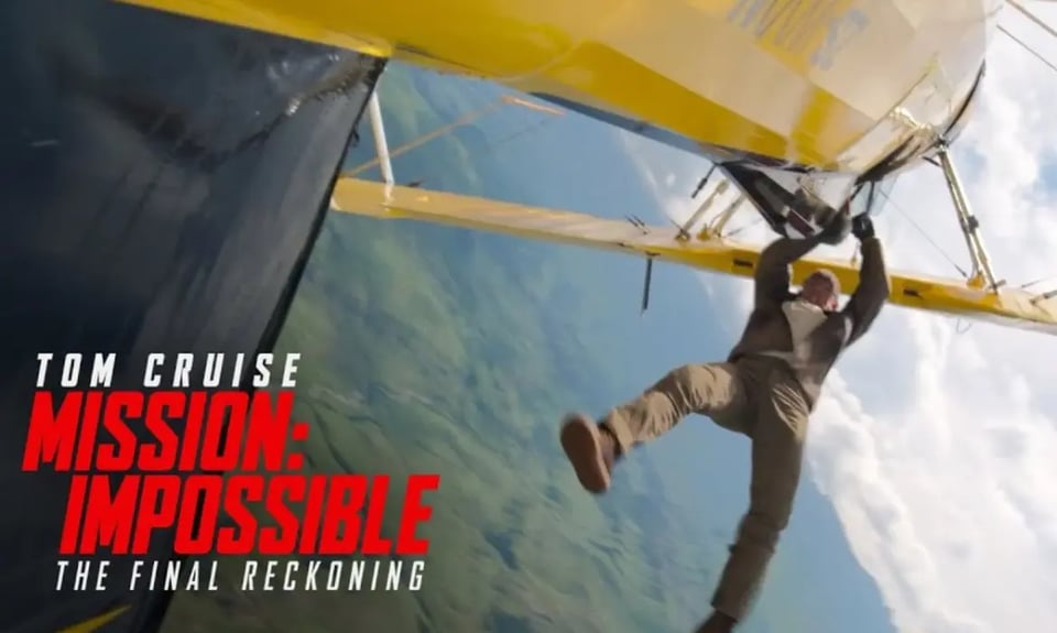 A person clings to the side of a yellow and white biplane in mid-flight, suspended against a blue sky. The shot is taken from below, showing them hanging from the aircraft's door whilst wearing tan trousers and boots. Text overlay reads "TOM CRUISE MISSION: IMPOSSIBLE THE FINAL RECKONING" in white and red letters.