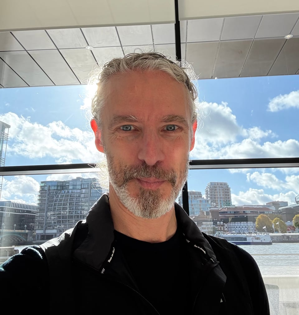 Selfie of white man with trim white beard, sunlight & the Thames behind him. He’s wearing a black pullover and a dark running vest, and has a smile that makes you want to know him better.