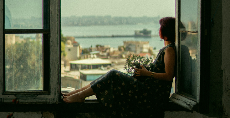 this is a photo of a person with chin length hair wearing a dress and holding a plant sitting inside a large window frame and looking out towards a harbour. This is the image that is on the front of the Free Window of Capacity resource.