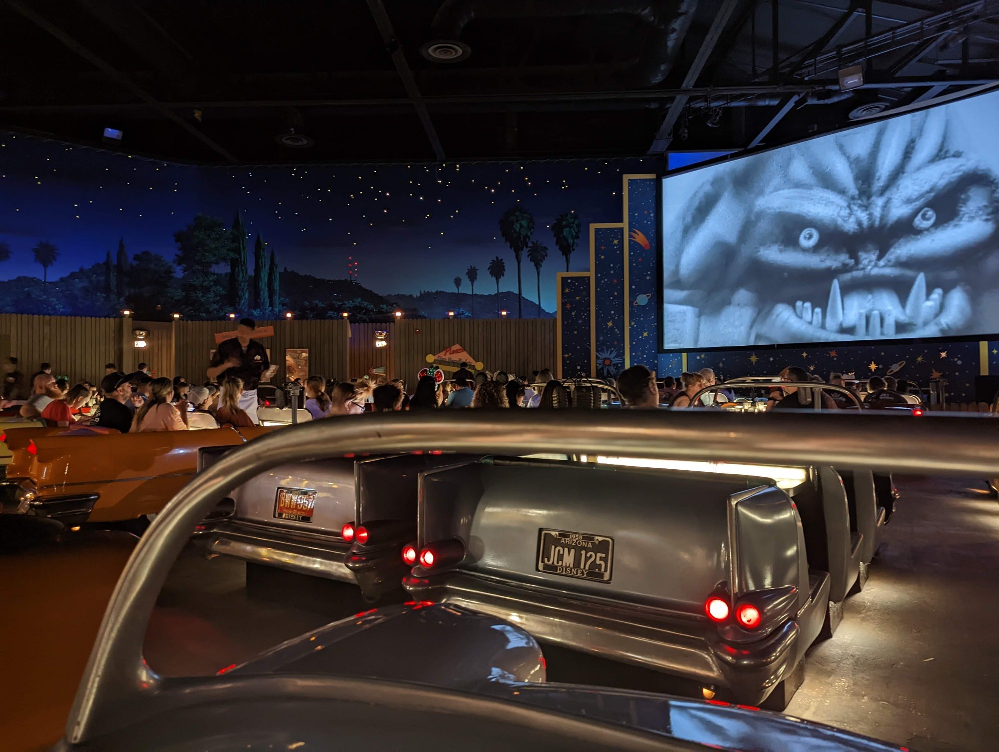 The interior of the Sci-Fi Dine In restaurant, styled to look like a drive-in movie theater at night. People sit in many pastel, 50s-style cars facing a big movie screen, which has a black-and-white image of a monster on it