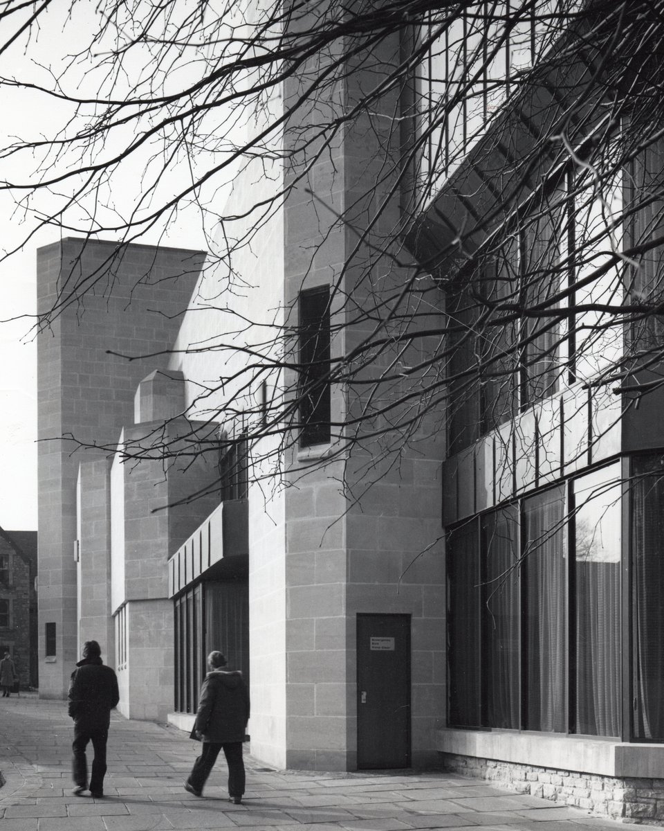 A large building of pale ashlar stone with a couple of people walking alongside it. The building is made up of several stone masses with cut-off corners. There's also a two-storey glass curtain wall. Above that, vertical seam lead cladding covers a jutting out block.