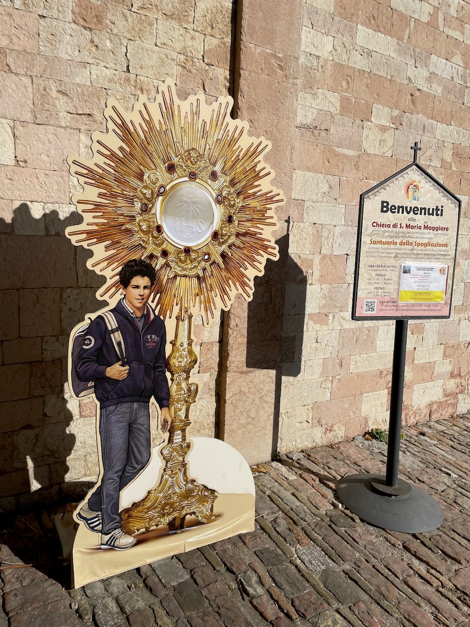 A cut-out image of Carlo Acutis, looking like any other Italian teen in a track jacket and with a bookbag, outside a medieval church in Assisi