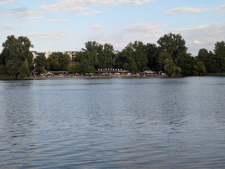A blue lake reflects a partly cloudy sky. Across the lake is a public beach with a lot of green, leafy trees. There is text on top of the facilities that reads WEISSENSEE, which is the name of the lake.