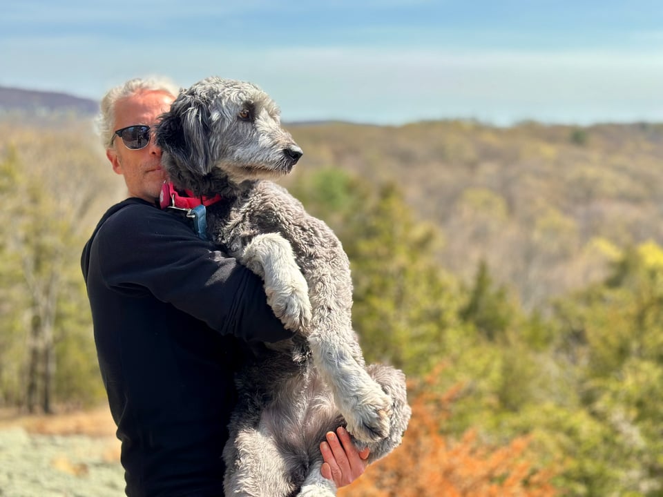 photo of white man in black hoodie & sunglasses holding up a 50+-lb. grey aussiedoodle, with woods and hills in the background
