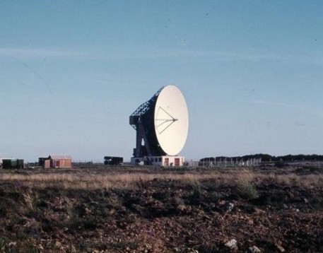 A grainy photo of a large parabolic dish, pointing horizontally. It's on a scrubby bit of moorland and against a faded blue sky.