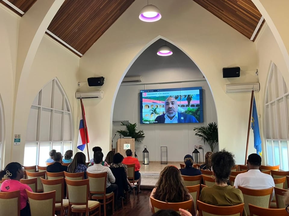 A picture of the event kick off (in the Aula of the University of Aruba), with the rector of the University of St. Martin on the screen and attendees listening and looking at the screen.