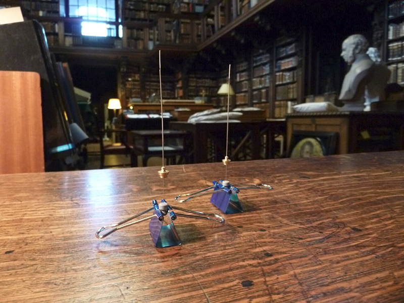 A close up picture of two electronic devices that are sitting on a wooden table in St Paul's cathedral. Each consist of thin metal pin -- perhaps 4 inches long -- each with a small circular object attached one third of the way up the pin. Each pin is being held upright in a vertical position by a document clip. In the background you can see desks and shelves of books and a white bust of an old man