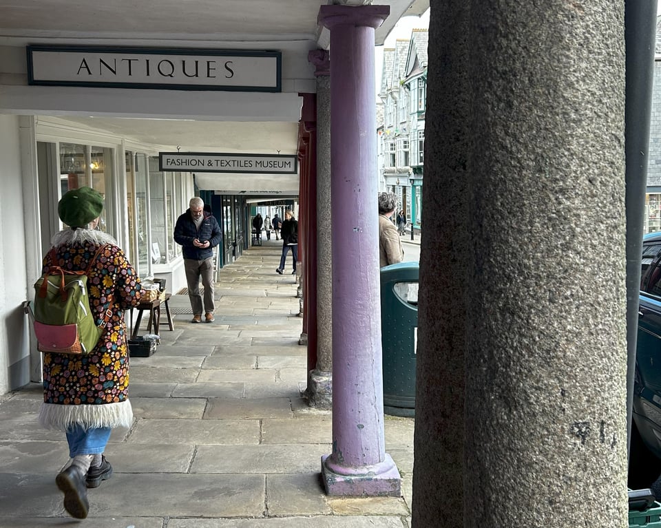 Shops under a colonnade of pillars that are supporting an overhanging first floor. Some pillars are stone, others are painted wood.