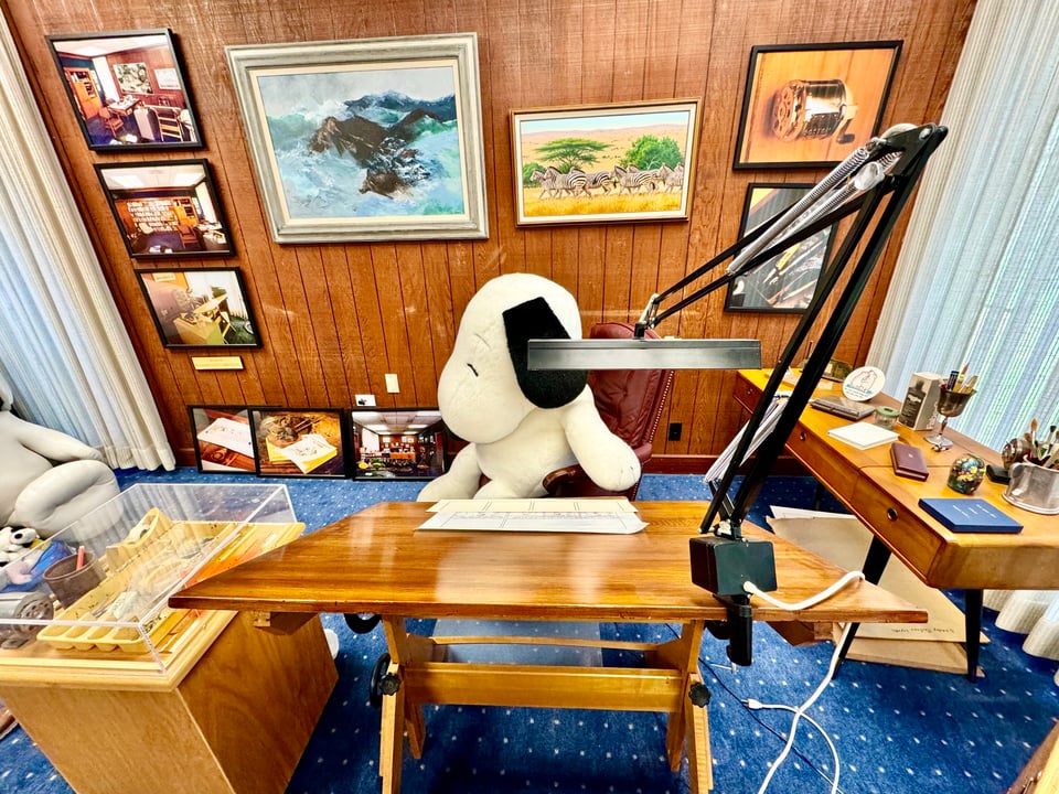 Photograph of Charles M. Schulz long-time studio with an oversized Snoopy plush animal sitting in his chair in front of a drawing board. Paintings and photographs hang on the wall behind him. The photo is taken from in front of the desk looking towards the seated party.