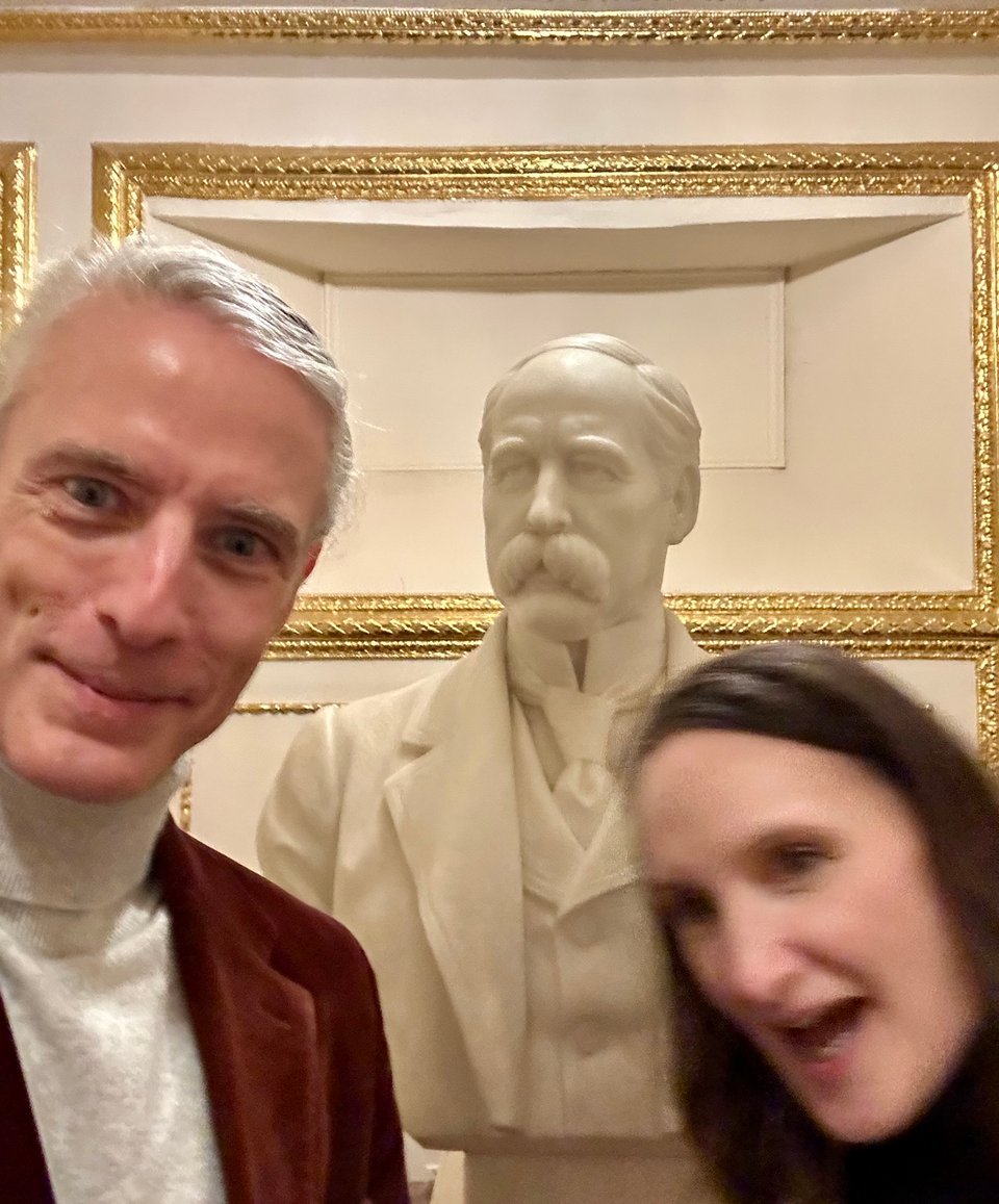 Selfie of white man in burgundy suit and light gray turtleneck, standing beside a white marble bust of a Gilded Age-era guy. A woman with dark hair is photobombing the shot from the right side of the frame